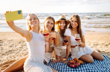 Neşeli kız arkadaşlar sahilde rahatlar, telefonda selfie çeker ve bardaktan içki içerler. Dört kadın deniz kıyısında pikniğin tadını çıkarıyor. Dostluk kavramı, kutlama.