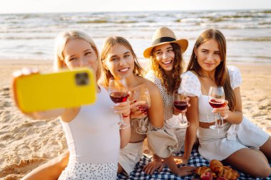 Neşeli kız arkadaşlar sahilde rahatlar, telefonda selfie çeker ve bardaktan içki içerler. Dört kadın deniz kıyısında pikniğin tadını çıkarıyor. Dostluk kavramı, kutlama.