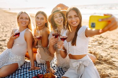 Neşeli kız arkadaşlar sahilde rahatlar, telefonda selfie çeker ve bardaktan içki içerler. Dört kadın deniz kıyısında pikniğin tadını çıkarıyor. Dostluk kavramı, kutlama.