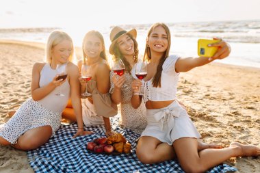 Neşeli kız arkadaşlar sahilde rahatlar, telefonda selfie çeker ve bardaktan içki içerler. Dört kadın deniz kıyısında pikniğin tadını çıkarıyor. Dostluk kavramı, kutlama.