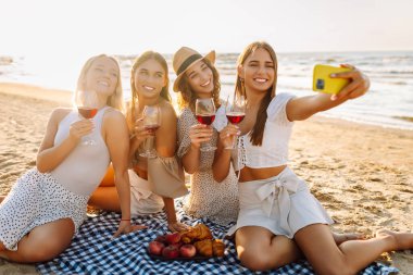 Neşeli kız arkadaşlar sahilde rahatlar, telefonda selfie çeker ve bardaktan içki içerler. Dört kadın deniz kıyısında pikniğin tadını çıkarıyor. Dostluk kavramı, kutlama.