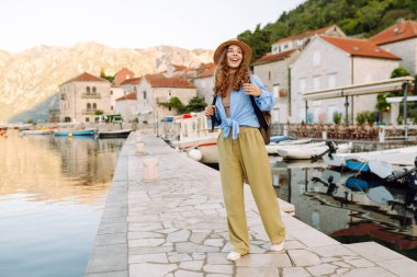 Şapkalı ve sırt çantalı güzel bir bayan gezgin gün batımında deniz manzarasının tadını çıkarıyor. Sırt çantalı genç bir kadın körfezin manzarasına hayran. Seyahat ve turizm konsepti. Etkin yaşam biçimi.