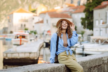 Şapkalı ve sırt çantalı güzel bir bayan gezgin gün batımında deniz manzarasının tadını çıkarıyor. Sırt çantalı genç bir kadın körfezin manzarasına hayran. Seyahat ve turizm konsepti. Etkin yaşam biçimi.