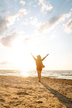 Deniz kıyısında günbatımının tadını çıkaran mutlu kadının arka görüntüsü. Kumsalda yürüyen ve özgürlüğü hisseden şapkalı güzel bir turist. Seyahat, eğlence konsepti. Etkin yaşam biçimi.