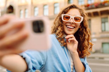 Güneş gözlüklü şık bir kadın güneşli şehir sokaklarında selfie çekiyor. Genç bir kadın gün batımında eğleniyor ve blog yazıyor. Blogculuk, turizm konsepti.