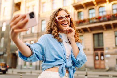 Güneş gözlüklü şık bir kadın güneşli şehir sokaklarında selfie çekiyor. Genç bir kadın gün batımında eğleniyor ve blog yazıyor. Blogculuk, turizm konsepti.
