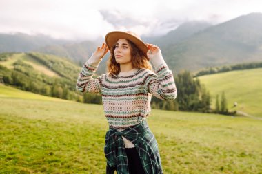 Şapkalı kadın gezgin yeşil tepede durmuş, eğleniyor ve dağ manzarasının tadını çıkarıyor. Rahat giysiler içindeki genç kadın dışarıda özgürlüğünü hissediyor. Macera, yürüyüş ve tatil konsepti.