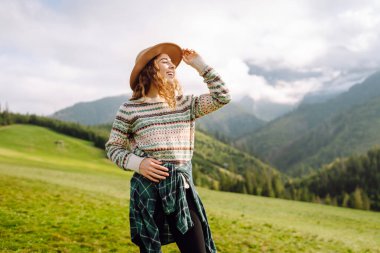 Şapkalı kadın gezgin yeşil tepede durmuş, eğleniyor ve dağ manzarasının tadını çıkarıyor. Rahat giysiler içindeki genç kadın dışarıda özgürlüğünü hissediyor. Macera, yürüyüş ve tatil konsepti.