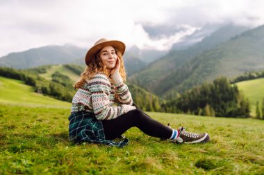 Şapkalı kadın gezgin yeşil tepede durmuş, eğleniyor ve dağ manzarasının tadını çıkarıyor. Rahat giysiler içindeki genç kadın dışarıda özgürlüğünü hissediyor. Macera, yürüyüş ve tatil konsepti.
