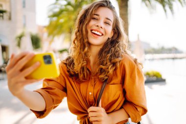 Şirin bir turist telefonuyla güneşli bir setin arka planına selfie çekiyor. Telefonu olan güzel bir kadın gün batımında yürüyüş ve blog yapmaktan hoşlanıyor. Turizm, blog konsepti.