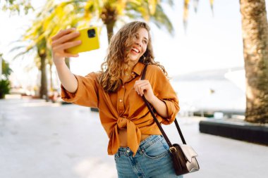 Şirin bir turist telefonuyla güneşli bir setin arka planına selfie çekiyor. Telefonu olan güzel bir kadın gün batımında yürüyüş ve blog yapmaktan hoşlanıyor. Turizm, blog konsepti.