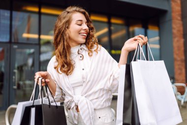 Alışveriş merkezinin dışındaki havanın tadını çıkaran neşeli bir kadın. Açık havadan alışveriş yapan genç bir turist. Aktif yaşam tarzı. İndirim ve tüketim kavramı.