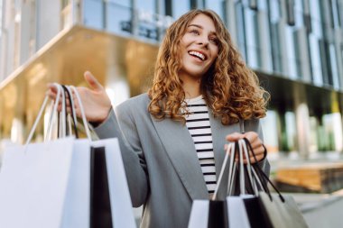 Günbatımında modern bir alışveriş merkezinin yakınında alışveriş torbalarıyla şık bir kadın. Alışverişten sonra yürüyüş yapan güzel bir kadın. Tüketim, tarz ve moda kavramı.