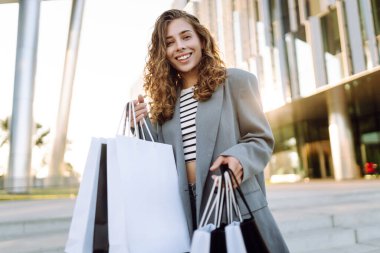 Günbatımında modern bir alışveriş merkezinin yakınında alışveriş torbalarıyla şık bir kadın. Alışverişten sonra yürüyüş yapan güzel bir kadın. Tüketim, tarz ve moda kavramı.