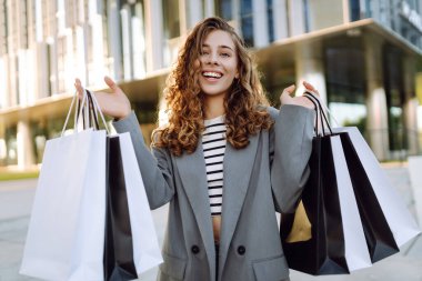 Günbatımında modern bir alışveriş merkezinin yakınında alışveriş torbalarıyla şık bir kadın. Alışverişten sonra yürüyüş yapan güzel bir kadın. Tüketim, tarz ve moda kavramı.