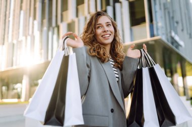 Günbatımında modern bir alışveriş merkezinin yakınında alışveriş torbalarıyla şık bir kadın. Alışverişten sonra yürüyüş yapan güzel bir kadın. Tüketim, tarz ve moda kavramı.