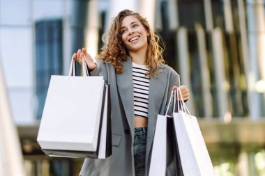 Günbatımında modern bir alışveriş merkezinin yakınında alışveriş torbalarıyla şık bir kadın. Alışverişten sonra yürüyüş yapan güzel bir kadın. Tüketim, tarz ve moda kavramı.