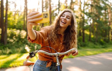 Güzel bir kadın güneşli bir parkta bisiklet sürerken selfie çeker. Genç bir kadın günbatımında bisiklet sürmekten hoşlanıyor. Etkin yaşam biçimi.