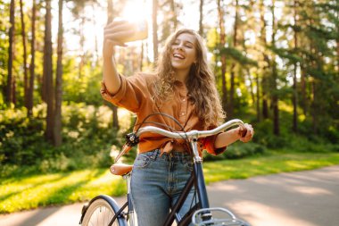 Güzel bir kadın güneşli bir parkta bisiklet sürerken selfie çeker. Genç bir kadın günbatımında bisiklet sürmekten hoşlanıyor. Etkin yaşam biçimi.