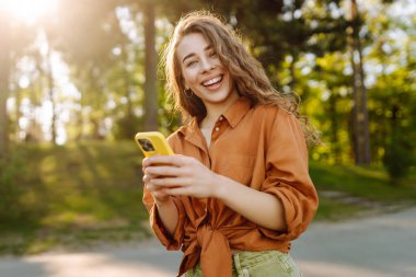 Akıllı telefonlu neşeli kadın portresi. Sunny Park 'ta mesaj atıp blog yazıyor. Dışarıda dinlenen ve elinde telefonla mesajlaşan genç bir kadın. Bloglama ve teknoloji kavramı.