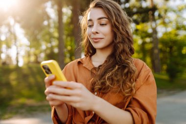 Akıllı telefonlu neşeli kadın portresi. Sunny Park 'ta mesaj atıp blog yazıyor. Dışarıda dinlenen ve elinde telefonla mesajlaşan genç bir kadın. Bloglama ve teknoloji kavramı.