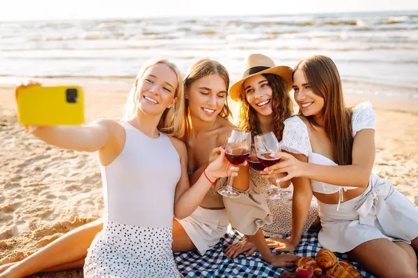 Neşeli kız arkadaşlar sahilde rahatlar, telefonda selfie çeker ve bardaktan içki içerler. Dört kadın deniz kıyısında pikniğin tadını çıkarıyor. Dostluk kavramı, kutlama.