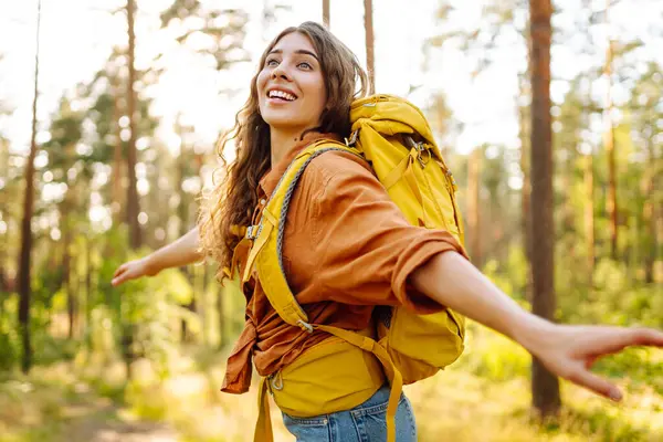Sarı bir sırt çantasıyla genç bir bayan gezgin özgürlüğü hissediyor ve ormanda günbatımının tadını çıkarıyor. Güzel bir kadın eğleniyor ve yürüyüş parkurunda yürüyor. Yürüyüş ve özgürlük kavramı.