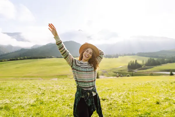 Şapkalı kadın gezgin yeşil tepede durmuş, eğleniyor ve dağ manzarasının tadını çıkarıyor. Rahat giysiler içindeki genç kadın dışarıda özgürlüğünü hissediyor. Macera, yürüyüş ve tatil konsepti.