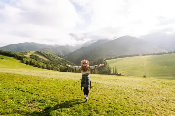Şapkalı kadın gezgin yeşil tepede durmuş, eğleniyor ve dağ manzarasının tadını çıkarıyor. Rahat giysiler içindeki genç kadın dışarıda özgürlüğünü hissediyor. Macera, yürüyüş ve tatil konsepti.