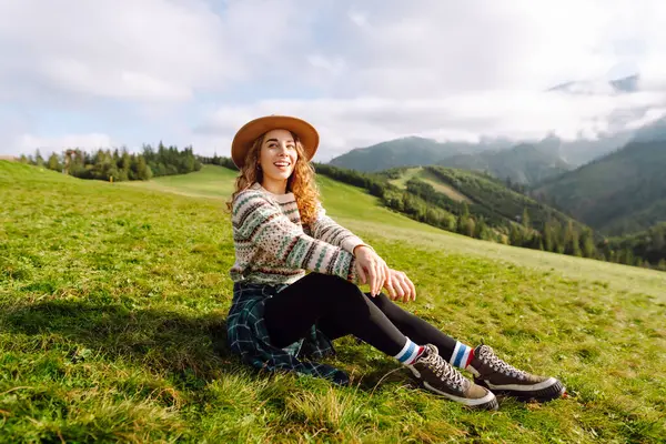 Şapkalı kadın gezgin yeşil tepede durmuş, eğleniyor ve dağ manzarasının tadını çıkarıyor. Rahat giysiler içindeki genç kadın dışarıda özgürlüğünü hissediyor. Macera, yürüyüş ve tatil konsepti.