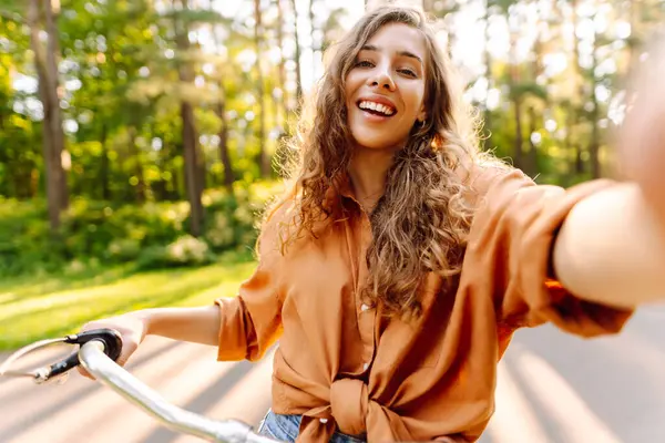 Güzel bir kadın güneşli bir parkta bisiklet sürerken selfie çeker. Genç bir kadın günbatımında bisiklet sürmekten hoşlanıyor. Etkin yaşam biçimi.