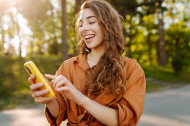 Akıllı telefonlu neşeli kadın portresi. Sunny Park 'ta mesaj atıp blog yazıyor. Dışarıda dinlenen ve elinde telefonla mesajlaşan genç bir kadın. Bloglama ve teknoloji kavramı.