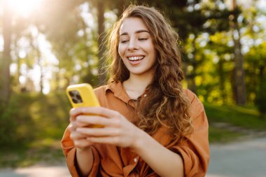 Akıllı telefonlu neşeli kadın portresi. Sunny Park 'ta mesaj atıp blog yazıyor. Dışarıda dinlenen ve elinde telefonla mesajlaşan genç bir kadın. Bloglama ve teknoloji kavramı.