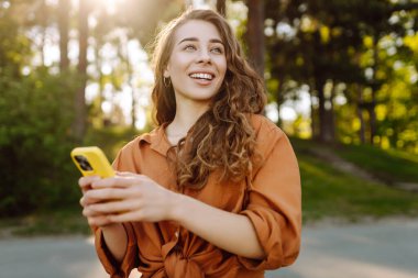 Akıllı telefonlu neşeli kadın portresi. Sunny Park 'ta mesaj atıp blog yazıyor. Dışarıda dinlenen ve elinde telefonla mesajlaşan genç bir kadın. Bloglama ve teknoloji kavramı.