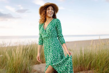 Hasır çanta ve şapkayla kumsalda yürüyen gülümseyen kadın. Genç turist denizde gün batımının tadını çıkarıyor ve dışarıda eğleniyor. Hafta sonu konsepti.