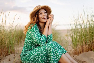 Hasır çanta ve şapkayla kumsalda yürüyen gülümseyen kadın. Genç turist denizde gün batımının tadını çıkarıyor ve dışarıda eğleniyor. Hafta sonu konsepti.