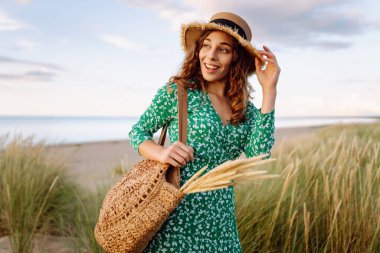 Hasır çanta ve şapkayla kumsalda yürüyen gülümseyen kadın. Genç turist denizde gün batımının tadını çıkarıyor ve dışarıda eğleniyor. Hafta sonu konsepti.
