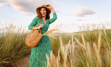 Hasır çanta ve şapkayla kumsalda yürüyen gülümseyen kadın. Genç turist denizde gün batımının tadını çıkarıyor ve dışarıda eğleniyor. Hafta sonu konsepti.