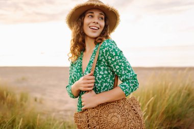 Hasır çanta ve şapkayla kumsalda yürüyen gülümseyen kadın. Genç turist denizde gün batımının tadını çıkarıyor ve dışarıda eğleniyor. Hafta sonu konsepti.