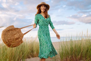 Hasır çanta ve şapkayla kumsalda yürüyen gülümseyen kadın. Genç turist denizde gün batımının tadını çıkarıyor ve dışarıda eğleniyor. Hafta sonu konsepti.