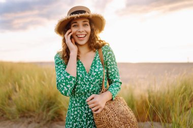 Hasır çanta ve şapkayla kumsalda yürüyen gülümseyen kadın. Genç turist denizde gün batımının tadını çıkarıyor ve dışarıda eğleniyor. Hafta sonu konsepti.