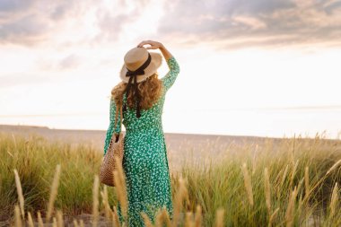 Hasır çanta ve şapkayla kumsalda yürüyen gülümseyen kadın. Genç turist denizde gün batımının tadını çıkarıyor ve dışarıda eğleniyor. Hafta sonu konsepti.