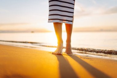 Gün batımında kumlu bir sahilde yürüyen genç bir kadının bacaklarının yakın çekimi. Güzel bir kadın sahilde yürürken günbatımının tadını çıkarıyor. Özgürlük kavramı.