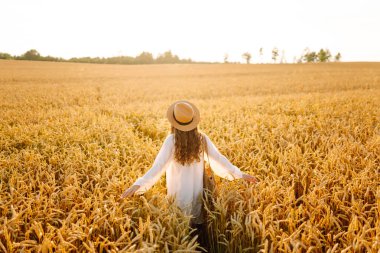 Beyaz elbiseli neşeli kadın altın buğday tarlasında günbatımının tadını çıkarıyor. Şapkalı güzel bir kadın dışarıda özgürce eğleniyor. Güzellik ve eğlence konsepti.
