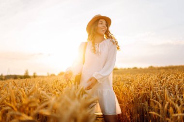 Beyaz elbiseli neşeli kadın altın buğday tarlasında günbatımının tadını çıkarıyor. Şapkalı güzel bir kadın dışarıda özgürce eğleniyor. Güzellik ve eğlence konsepti.