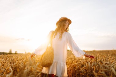 Beyaz elbiseli neşeli kadın altın buğday tarlasında günbatımının tadını çıkarıyor. Şapkalı güzel bir kadın dışarıda özgürce eğleniyor. Güzellik ve eğlence konsepti.