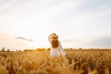 Beyaz elbiseli neşeli kadın altın buğday tarlasında günbatımının tadını çıkarıyor. Şapkalı güzel bir kadın dışarıda özgürce eğleniyor. Güzellik ve eğlence konsepti.
