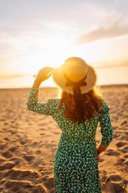 Gün batımı kumsalında saman şapkalı mutlu bir kadının portresi. Elbiseli genç bir kadın deniz manzarasından zevk alıyor, özgürlüğü hissediyor. Etkin yaşam biçimi