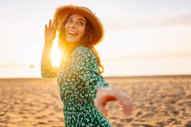 Gün batımı kumsalında saman şapkalı mutlu bir kadının portresi. Elbiseli genç bir kadın deniz manzarasından zevk alıyor, özgürlüğü hissediyor. Etkin yaşam biçimi