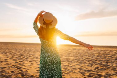 Gün batımı kumsalında saman şapkalı mutlu bir kadının portresi. Elbiseli genç bir kadın deniz manzarasından zevk alıyor, özgürlüğü hissediyor. Etkin yaşam biçimi